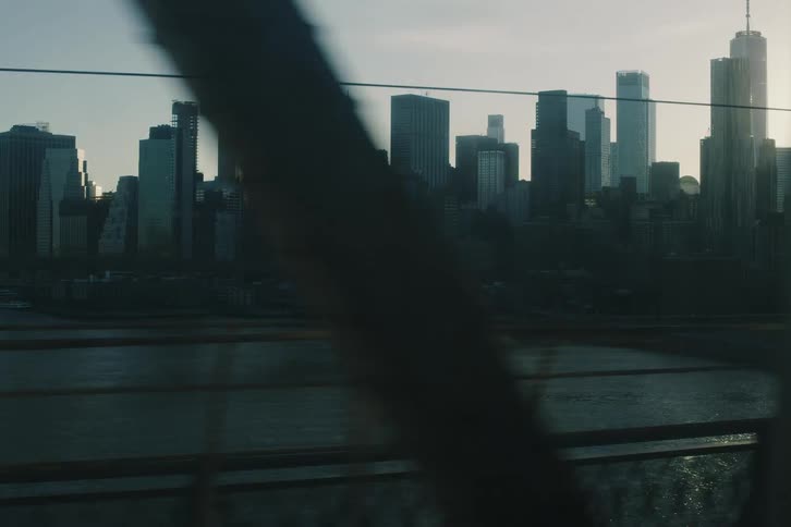 An atmospheric tracking shot of the New York City skyline at sunrise, with lens flare and the structure of a bridge passing in the foreground
