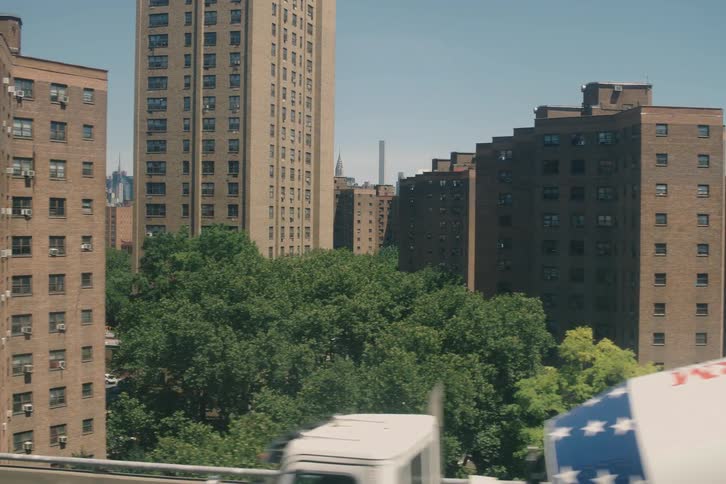 A tracking shot from a moving vehicle reveals the iconic New York City skyline behind residential apartment buildings on a sunny day