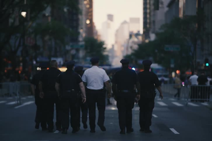 A group of police officers and their superior walk in slow motion down a barricaded city street at dusk