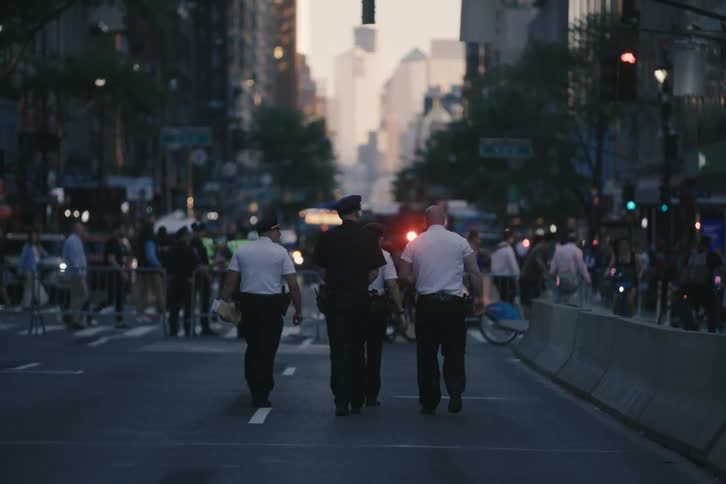 A group of police officers walk away from the camera down a crowded city street at dusk