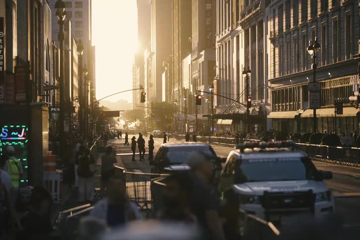 Crowds gather behind barricades on a sun-drenched city street guarded by police cars during a golden sunset