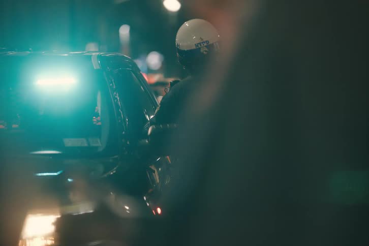 A New York City police officer stands by their patrol car at night, illuminated by flashing red and blue emergency lights