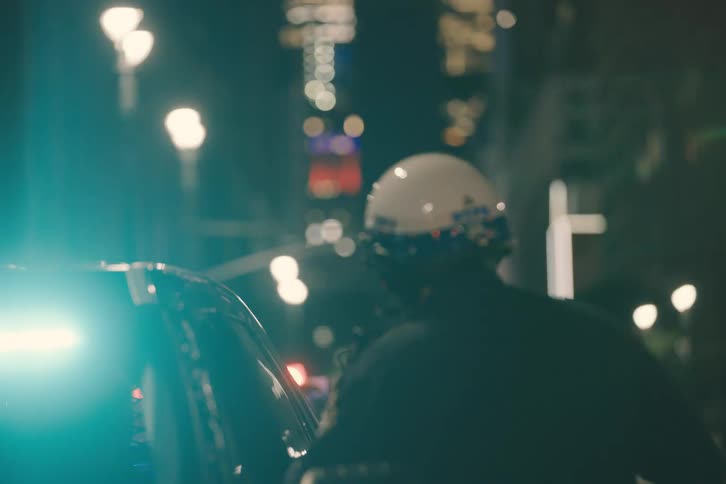 A New York City police officer stands by a vehicle at night as emergency lights flash and the city skyline looms in the background