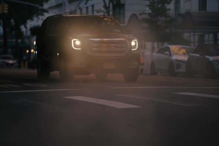 Cars drive through rising steam on a city street at dusk with police lights flashing in the background