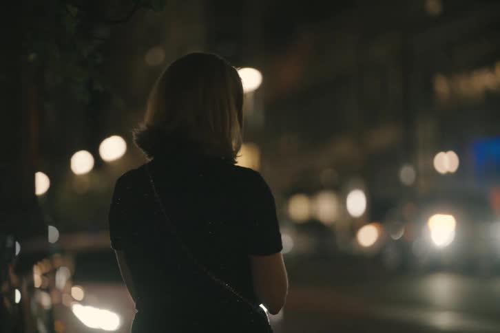Rack focus from a woman's silhouette to a bustling city street at night lined with luxury sports cars