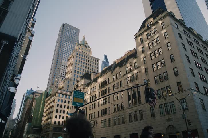 A low-angle view shows the sun hitting the side of the historic Bergdorf Goodman building on a busy street in New York City