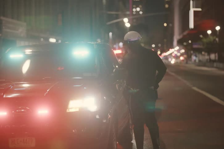 A police officer stands next to their patrol car with flashing lights on a busy city street at night