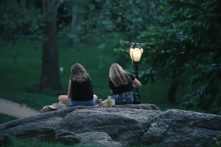 Two friends sit on a boulder and enjoy a peaceful evening in a city park next to a lit lamppost