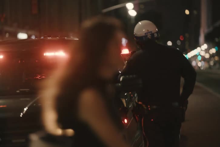 A New York City police officer stands watch by their patrol car on a bustling city street at night as the emergency lights flash