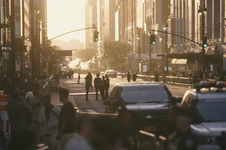 Police officers monitor a barricaded city street filled with waiting crowds during a hazy, golden hour sunset