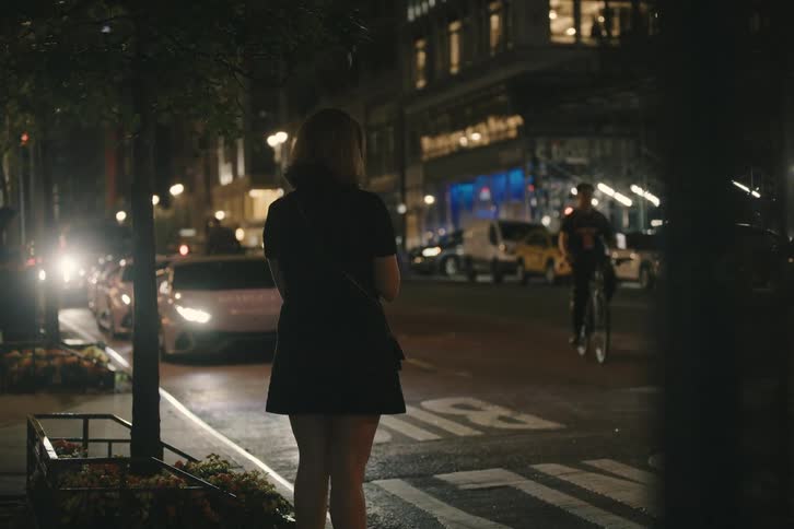 A woman in a black dress stands on a city sidewalk at night, watching the glowing headlights of a pink Lamborghini and other cars