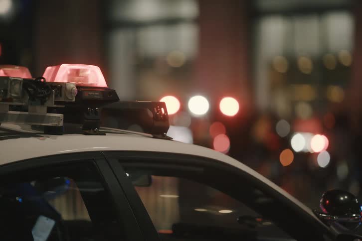 A close-up shot of a police car's flashing emergency lights on a city street at night, with a motorcade and pedestrians in the background