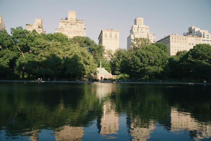A serene, static shot of New York City buildings reflected in the calm waters of a lake in Central Park on a sunny day