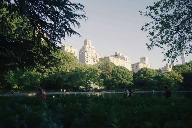 A slow pan reveals people enjoying a peaceful afternoon by the lake in Central Park with the iconic New York City skyline rising behind the trees