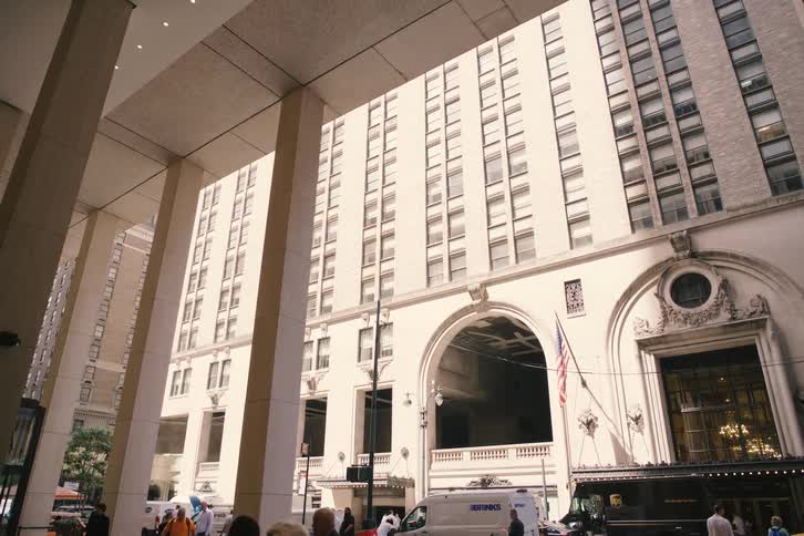 A low-angle tracking shot from behind large pillars reveals a grand building in a bustling city