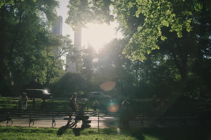 The camera reveals a sun-drenched scene of people walking and relaxing in a lush city park with the skyline visible in the background