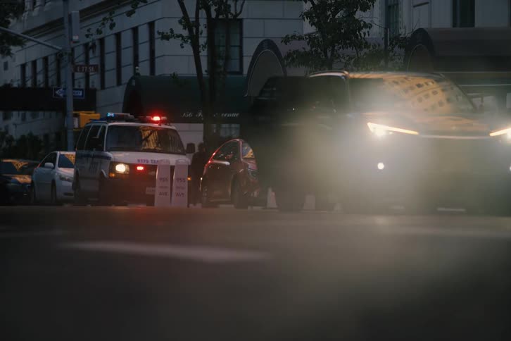 Steam rises from a New York City street at dusk as pedestrians cross in front of a police car with flashing emergency lights