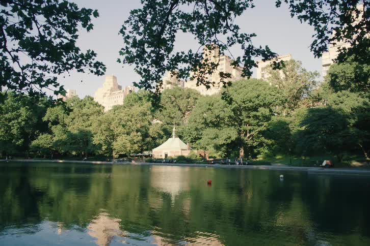 A serene view of the Kerbs Boathouse on Conservatory Water in New York City's Central Park, with lush green trees and city buildings reflected in the calm water