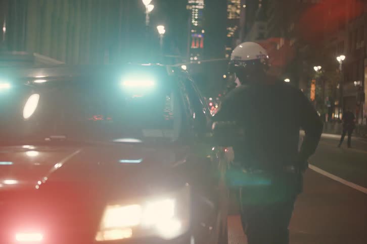 From behind, a police officer stands watch next to his cruiser as red and blue lights flash on a city street at night