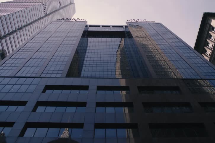A slow pull-back shot from a low angle shows the towering glass facade of a modern skyscraper reflecting the city skyline