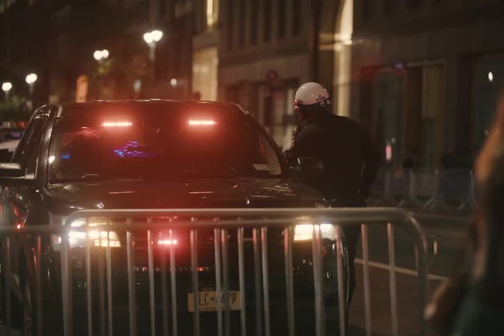 An NYPD officer stands guard by a police vehicle with flashing emergency lights on a city street at night