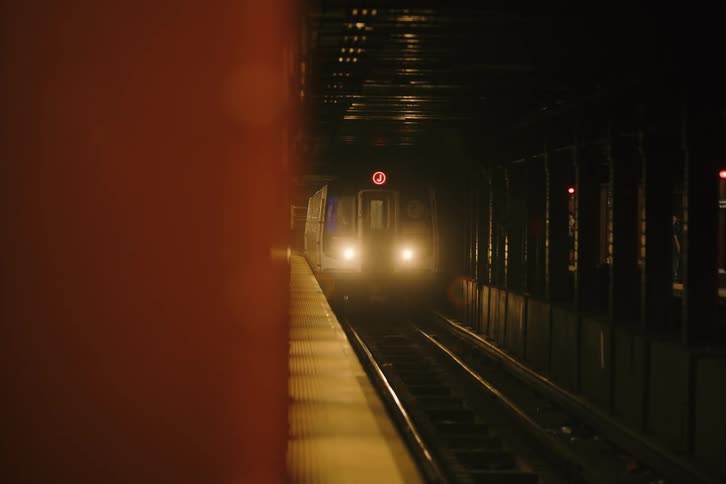 A New York City J train emerges from a dark tunnel, its bright headlights illuminating the tracks as it pulls into the station at night