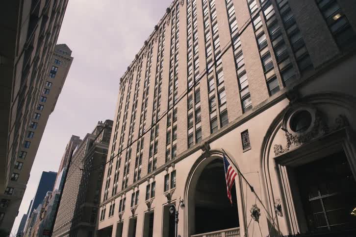 A slow, low-angle tracking shot moves towards a classic high-rise building on a New York City street with an American flag