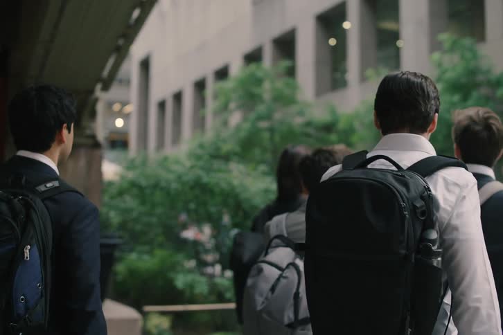A group of young professionals with backpacks walk in slow motion through the busy streets of New York City, starting at Pershing Square