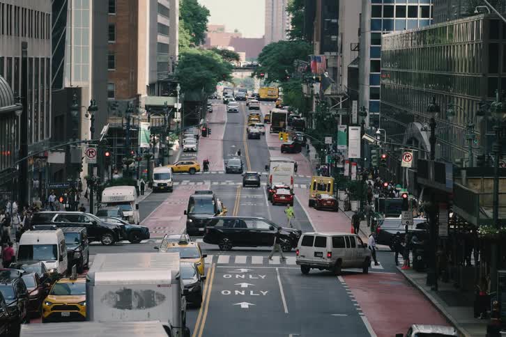 A high-angle static shot captures the bustling activity of a downtown Manhattan street, with a steady stream of traffic and pedestrians crossing at an intersection