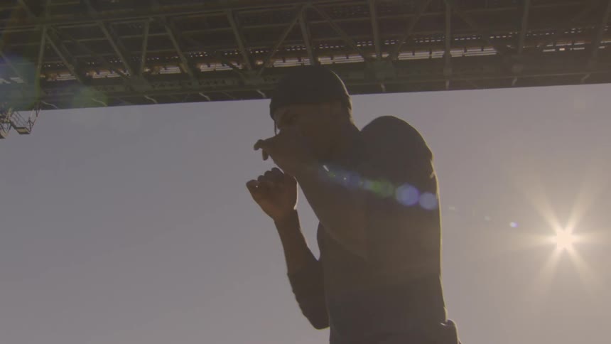 A man performs a slow-motion expressive dance beneath a large industrial bridge as the sun flares behind him
