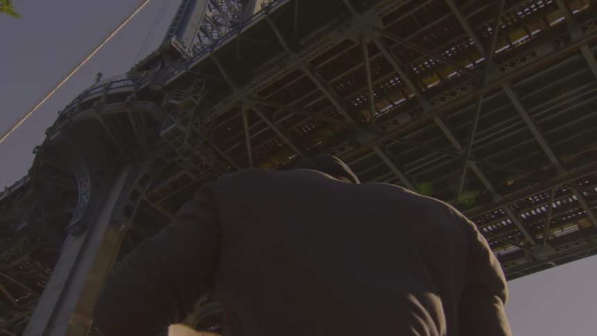 A young man wearing a beanie dances and contemplates in slow motion under the steel structure of a massive city bridge