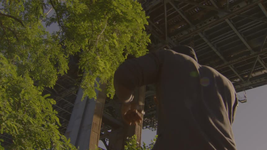 A low-angle slow-motion shot captures a man performing an interpretive dance under a large city bridge at golden hour