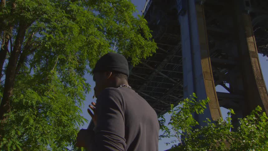 A man from behind smokes a cigarette in a park underneath a massive city bridge against a clear blue sky