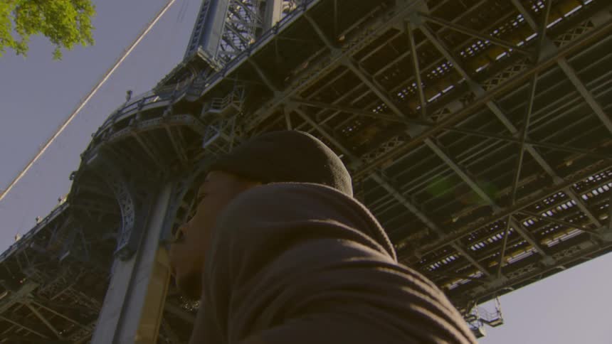 A thoughtful young man stands beneath a large city bridge, filmed from a low angle in slow motion as the camera pulls away