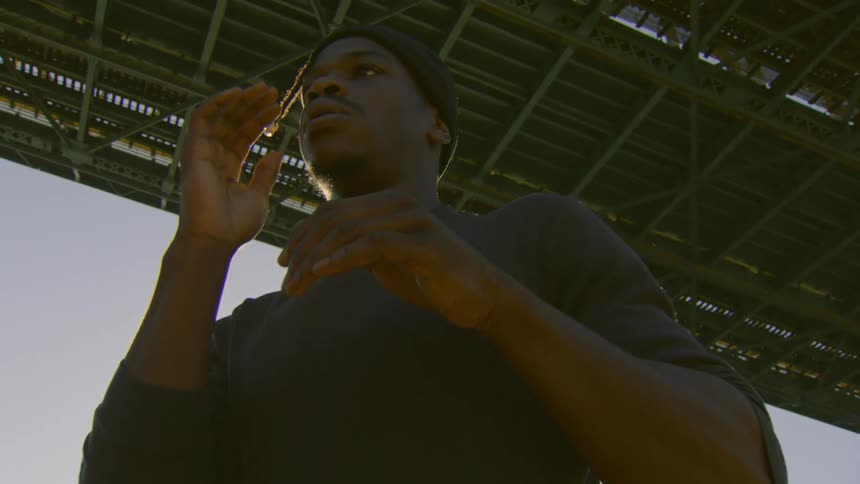 From a low angle, a focused young man performs a slow, expressive dance beneath a large industrial bridge as the sun sets