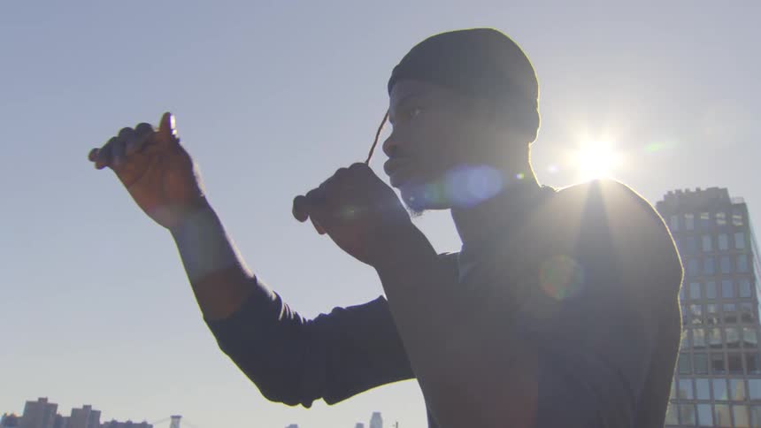 A determined young man warms up by stretching his arms in slow motion, backlit by the sun against a city skyline