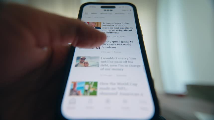 A close-up, low-angle shot of a person's thumb endlessly scrolling through a news feed on a smartphone