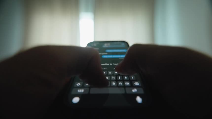 A close-up point-of-view shot of hands typing a text message conversation on a smartphone in a dimly lit room