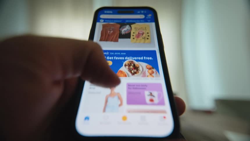 A close-up, point-of-view shot of a person scrolling through a retail shopping app on their smartphone, browsing various products like groceries, clothing, and home goods