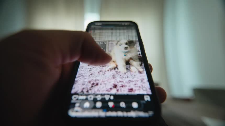 A close-up point of view shot of a person's hand scrolling through a social media feed on a smartphone in a dimly lit room
