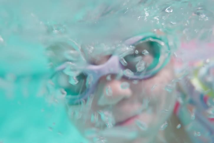 Slow motion underwater close-up of a little girl in goggles holding her breath and blowing bubbles in a swimming pool