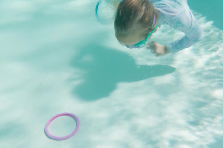 Playful little girl jumps into the pool and swims underwater to collect colorful diving rings in slow motion