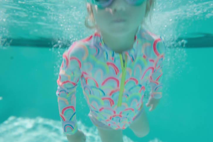 Young girl in colorful swimsuit jumps into a backyard pool and swims underwater toward the camera