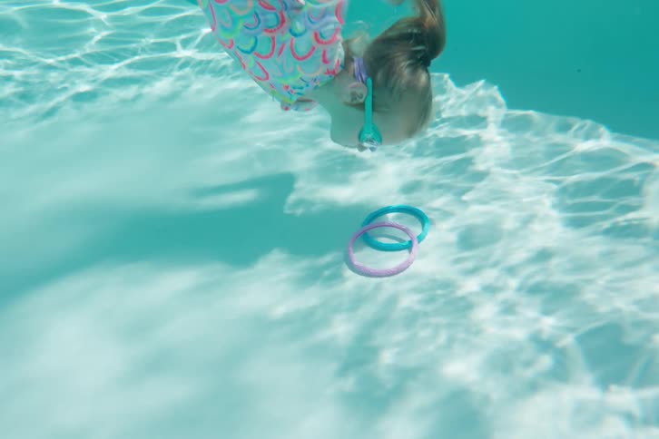 An underwater slow-motion shot shows a young girl in a colorful swimsuit and goggles diving to retrieve two rings from the bottom of a bright blue swimming pool