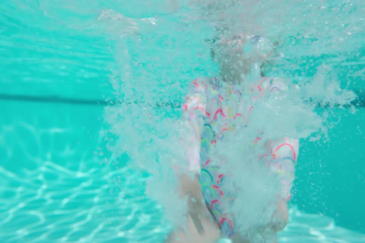 A little girl in a colorful swimsuit and goggles playfully swims underwater in a turquoise pool on a sunny day