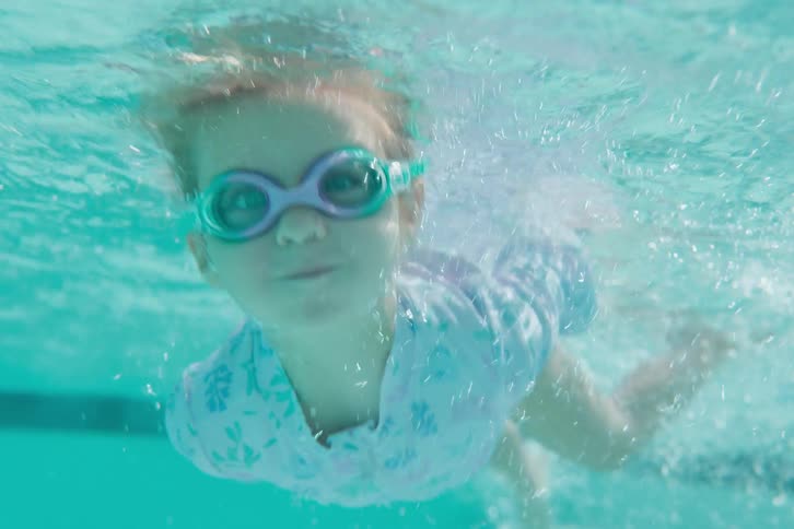 A young girl with goggles excitedly jumps into a swimming pool and swims underwater towards the camera in slow motion