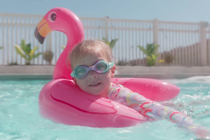 A happy toddler girl wearing goggles smiles as she playfully paddles on an inflatable pink flamingo float in a sunny backyard swimming pool in slow motion