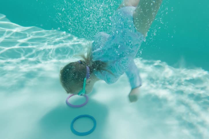A young girl in goggles jumps into a pool and dives underwater in slow motion to retrieve colorful rings from the bottom