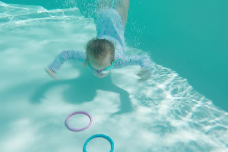 A little girl with goggles splashes into a pool and swims underwater to collect colorful diving rings from the bottom