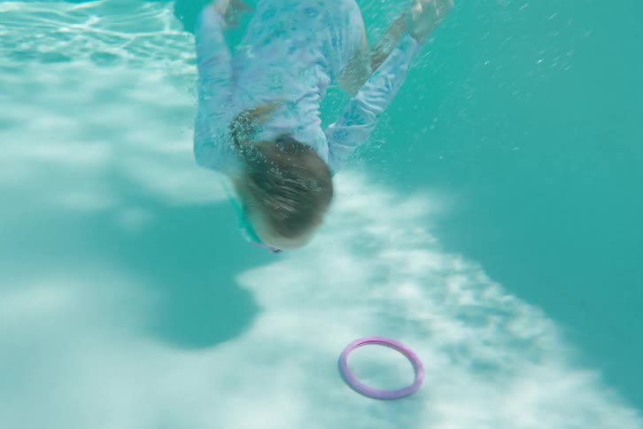 A young girl in goggles jumps into a swimming pool and dives underwater to retrieve a toy ring in slow motion